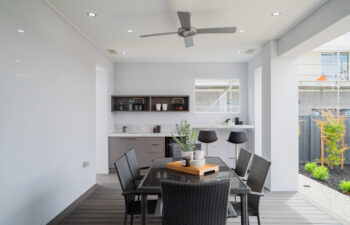 Modern covered alfresco area featuring a outdoor dining set, grey decking, ceiling fan, and built-in bar counter with stools and cabinetry.