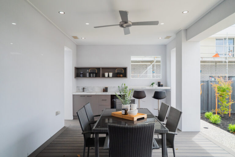 Modern covered alfresco area featuring a outdoor dining set, grey decking, ceiling fan, and built-in bar counter with stools and cabinetry.