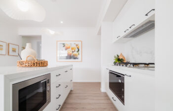 Modern white kitchen featuring a long island with a built-in microwave, marble-look counter and splashback, integrated appliances, and wavy pendant lights.