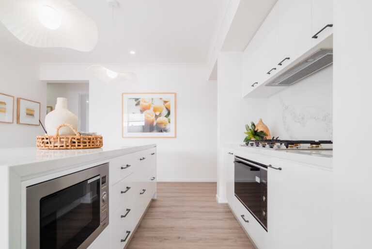 Modern white kitchen featuring a long island with a built-in microwave, marble-look counter and splashback, integrated appliances, and wavy pendant lights.