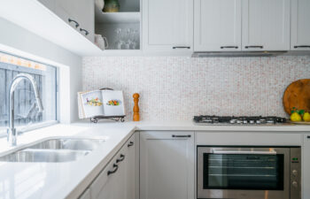 Modern kitchen corner featuring light grey shaker cabinetry, white counter, dual-bowl sink beneath a window, built-in oven, gas stovetop, and mosaic tile backsplash.
