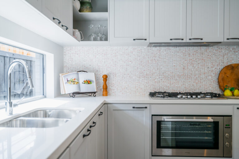 Modern kitchen corner featuring light grey shaker cabinetry, white counter, dual-bowl sink beneath a window, built-in oven, gas stovetop, and mosaic tile backsplash.