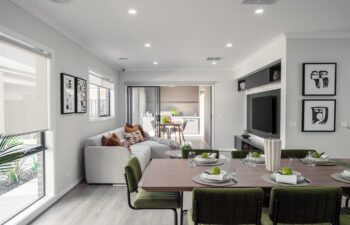 Modern open-plan living and dining area with olive green chairs around a timber table, a sectional sofa, media wall, and sliding doors leading to an outdoor alfresco space.