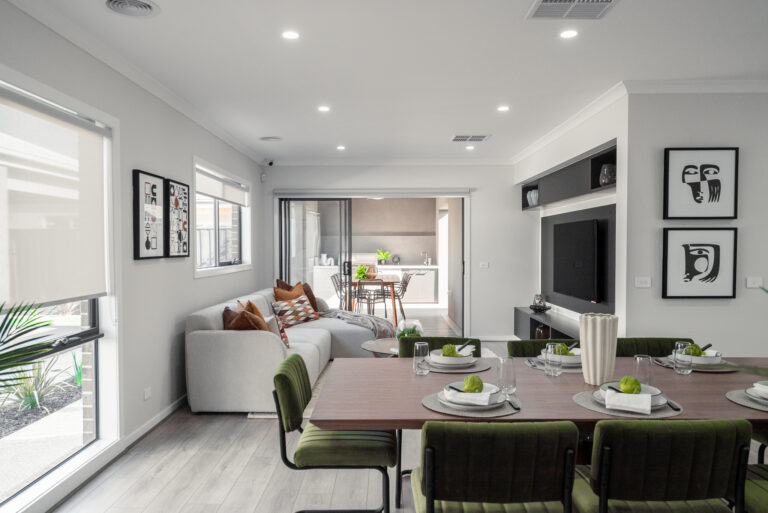 Modern open-plan living and dining area with olive green chairs around a timber table, a sectional sofa, media wall, and sliding doors leading to an outdoor alfresco space.