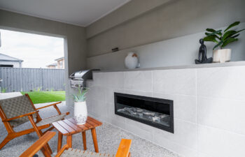 Modern covered outdoor alfresco area featuring a built-in linear fireplace, integrated barbecue grill, and wooden patio seating looking out toward a backyard.