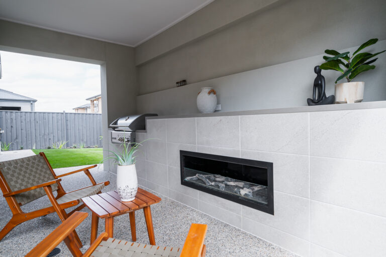 Modern covered outdoor alfresco area featuring a built-in linear fireplace, integrated barbecue grill, and wooden patio seating looking out toward a backyard.