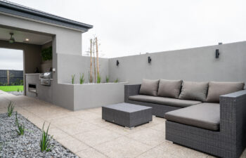 Modern outdoor alfresco design featuring built-in outdoor kitchen with BBQ, dark wicker lounge seating, aggregate paving, raised garden beds, and rendered privacy walls.