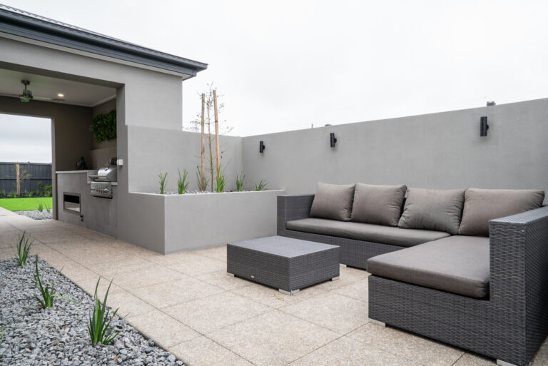 Modern outdoor alfresco design featuring built-in outdoor kitchen with BBQ, dark wicker lounge seating, aggregate paving, raised garden beds, and rendered privacy walls.