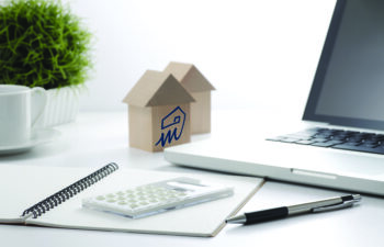 Home buyer and mortgage planning workspace with wooden model houses, laptop computer, calculator, notebook, and office accessories on a bright white desk.