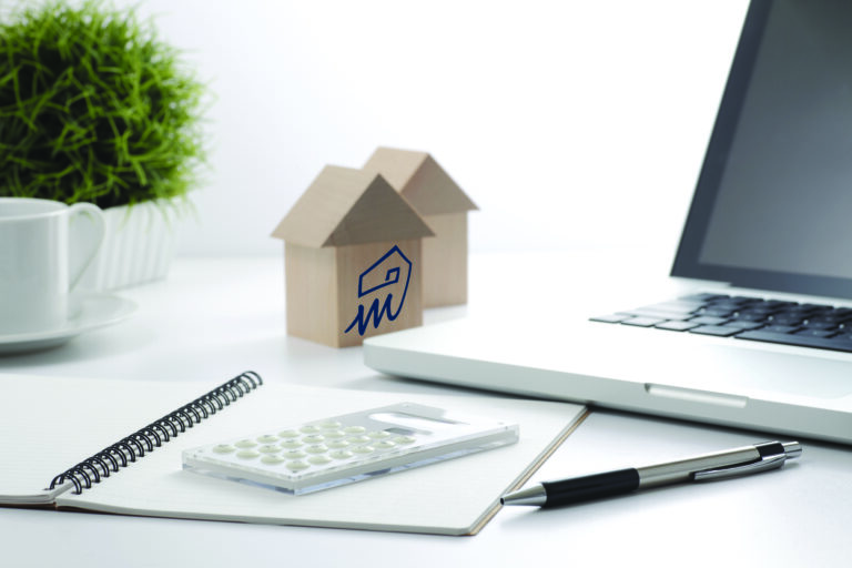 Home buyer and mortgage planning workspace with wooden model houses, laptop computer, calculator, notebook, and office accessories on a bright white desk.