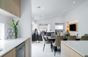 View from a modern kitchen looking into an open-plan dining and living area with white walls, tile flooring, and a recessed TV feature wall.