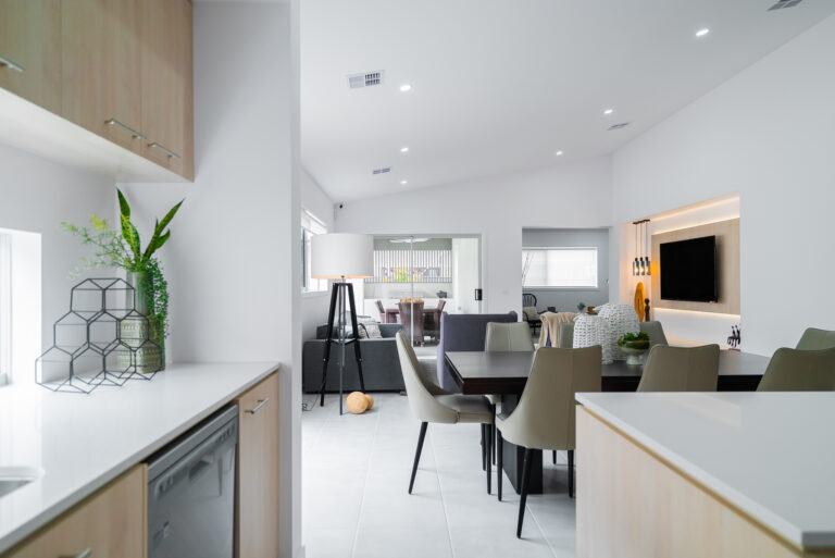 View from a modern kitchen looking into an open-plan dining and living area with white walls, tile flooring, and a recessed TV feature wall.
