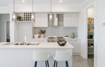 Bright modern kitchen featuring a white island bench with bar stools, patterned backsplash tiles, three glass pendant lights, and an open walk-in pantry.