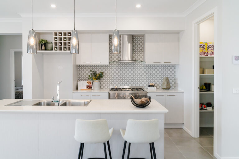 Bright modern kitchen featuring a white island bench with bar stools, patterned backsplash tiles, three glass pendant lights, and an open walk-in pantry.