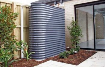 A slimline corrugated metal rainwater tank in dark grey installed along the side of a brick house, connected to roof downpipes.