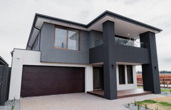 Contemporary two-story home facade with charcoal and off-white render, a covered upper balcony, dark timber garage door, and aggregate concrete driveway.