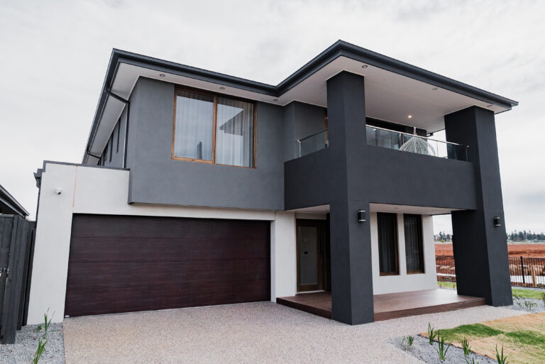 Contemporary two-story home facade with charcoal and off-white render, a covered upper balcony, dark timber garage door, and aggregate concrete driveway.