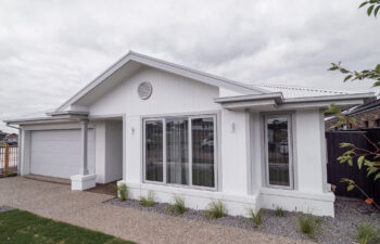 Single-story white coastal home featuring vertical cladding, grey window frames, a double garage, and exposed aggregate driveway.