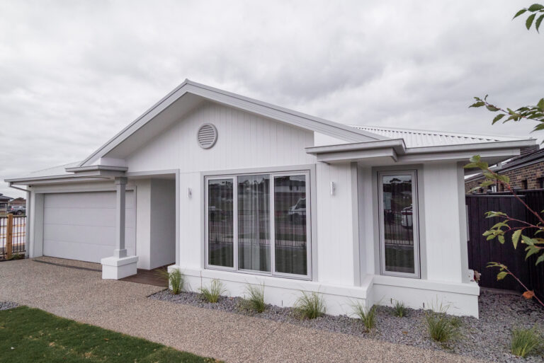 Single-story white coastal home featuring vertical cladding, grey window frames, a double garage, and exposed aggregate driveway.