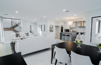 Open-plan living space featuring a dark dining set in the foreground, white sofas, a wooden staircase with black balusters, and a modern kitchen with a waterfall island.