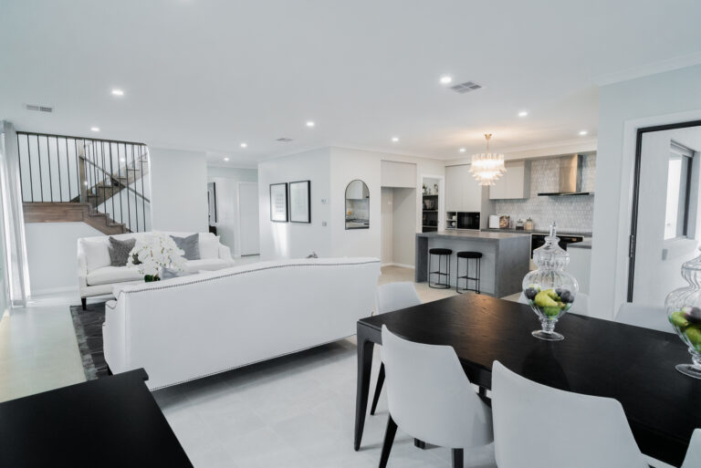 Open-plan living space featuring a dark dining set in the foreground, white sofas, a wooden staircase with black balusters, and a modern kitchen with a waterfall island.