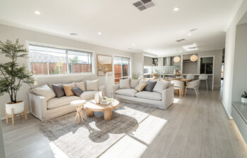 Spacious, light-filled open-plan living room with two beige sofas, a round wooden coffee table, grey area rug, and an adjoining kitchen and dining area.