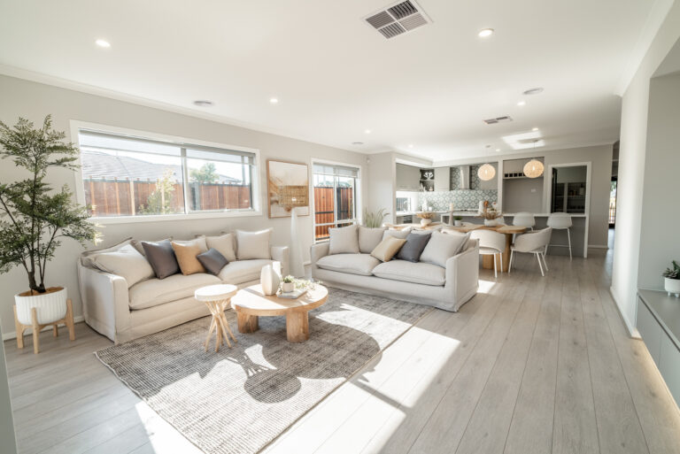 Spacious, light-filled open-plan living room with two beige sofas, a round wooden coffee table, grey area rug, and an adjoining kitchen and dining area.