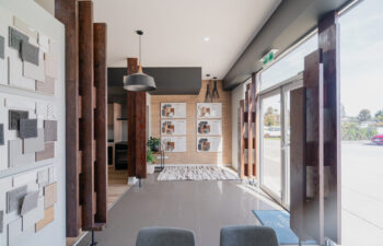 Modern interior design studio showroom featuring material sample boards, dark timber posts, tile flooring, and glass entry doors.