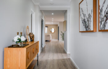 Bright modern hallway with light wood flooring, a wooden console table styled with flowers, framed botanical wall art, and a view leading into a living room.
