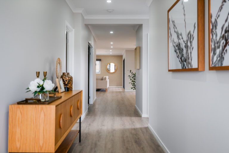 Bright modern hallway with light wood flooring, a wooden console table styled with flowers, framed botanical wall art, and a view leading into a living room.