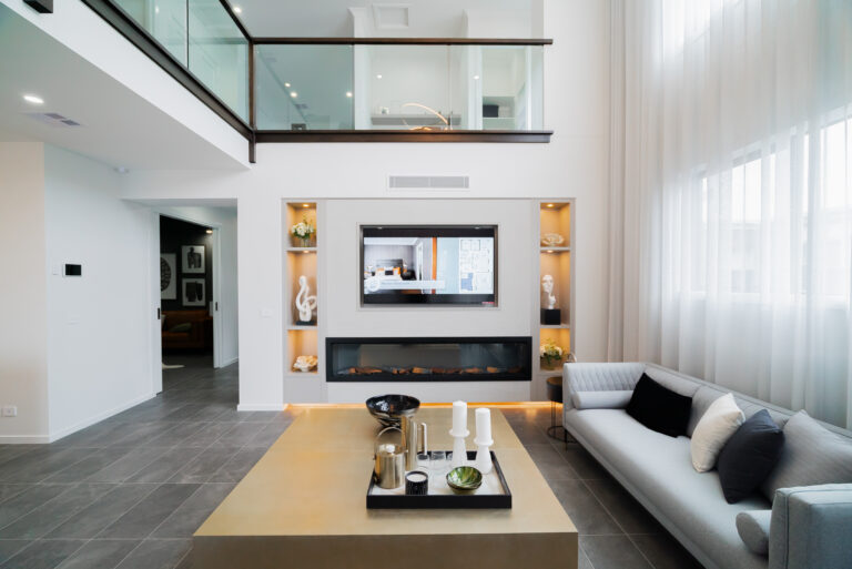Malvern 443 - Grand Atrium, Modern atrium-style living area featuring double-height ceilings, a second-story glass mezzanine, an integrated fireplace, and a large brass coffee table.