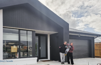 A family standing outside a modern single-story house with black trim, a garage, and a white interior doorway.